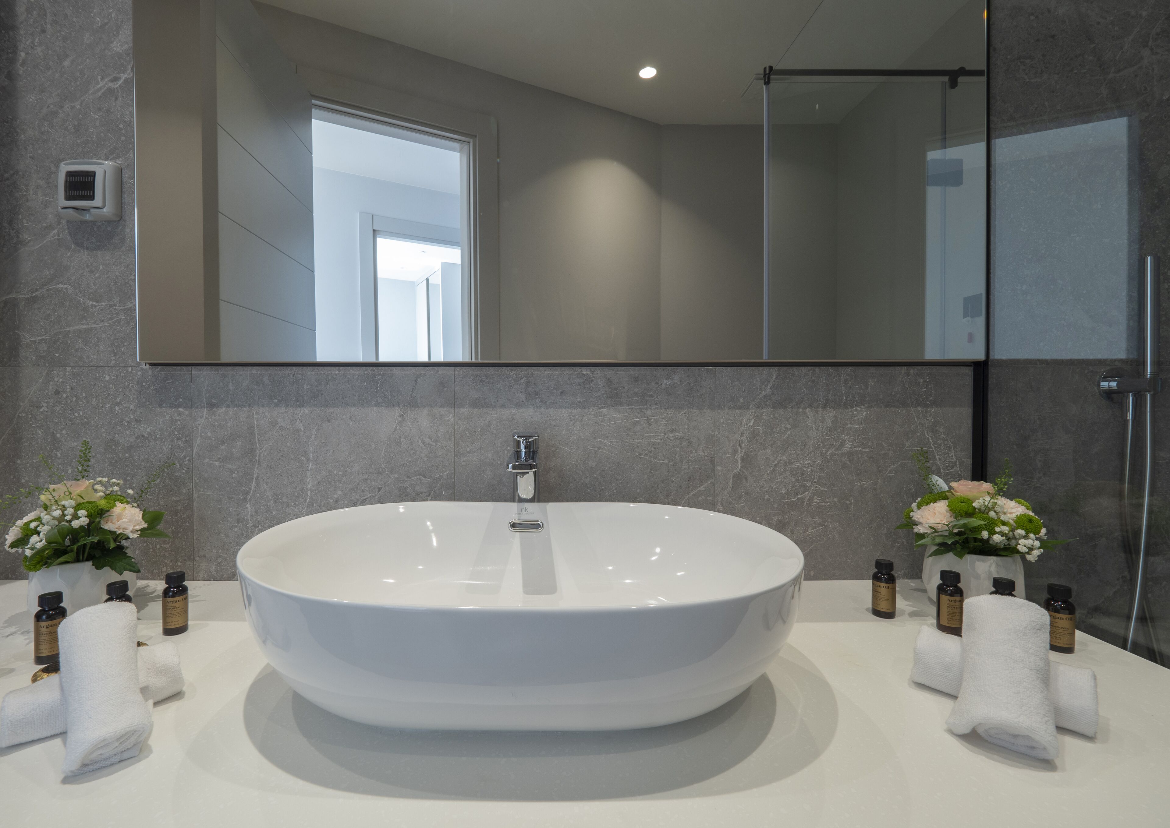 A modern bathroom with a large oval sink, a mirror above it, and two flower arrangements on the counter.