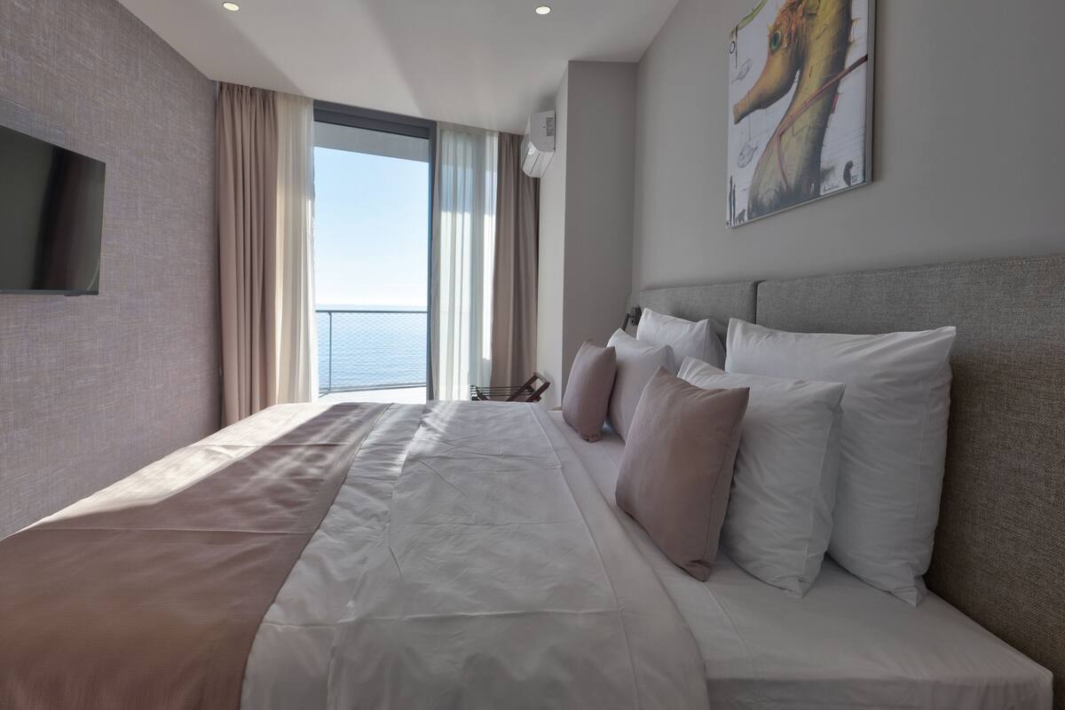 A modern hotel room with a large bed, a flat-screen TV, a view of the sea, and a painting on the wall.