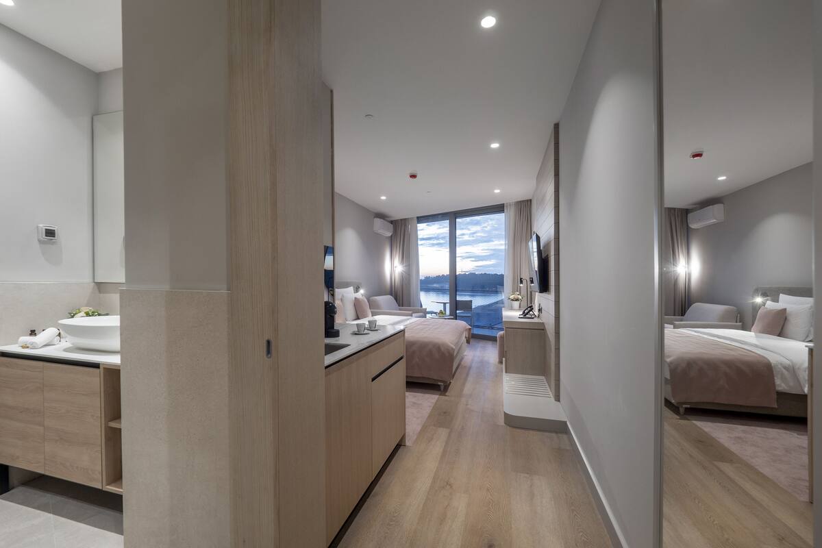 A modern hotel room with a large bed, a separate bathroom, a small kitchenette, and a view of the sea.