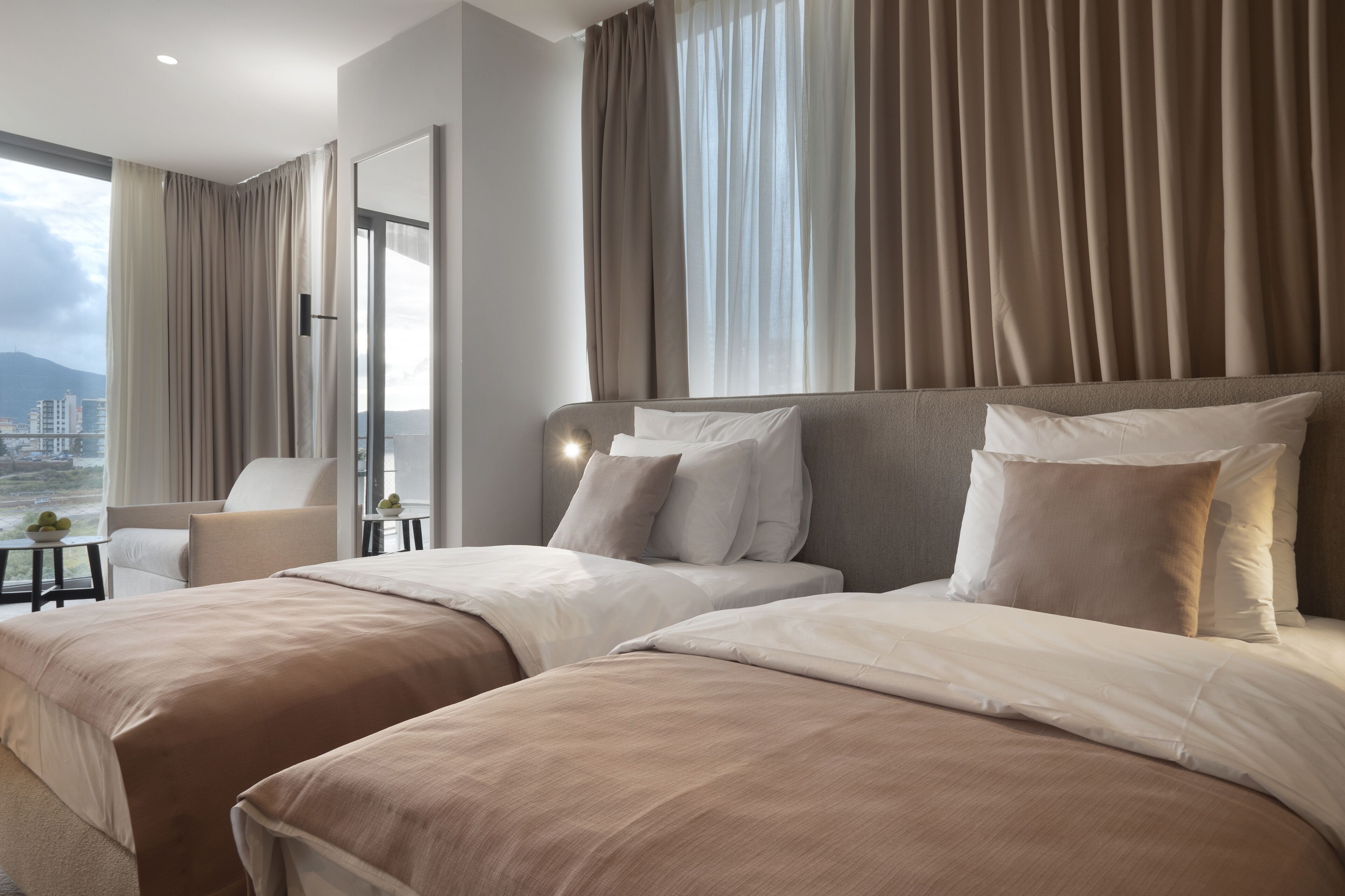 Et hotelværelse med to senge, et siddeområde med en stol, et lille bord med en lampe og et stort vindue med gardiner.