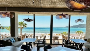 Un comedor interior con mesas y sillas, ventanas amplias con vista a la playa y al océano.