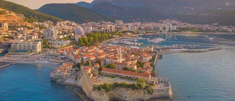Un paysage urbain côtier avec une imposante forteresse historique située sur une petite île, entourée de bâtiments résidentiels et d’un port de plaisance.