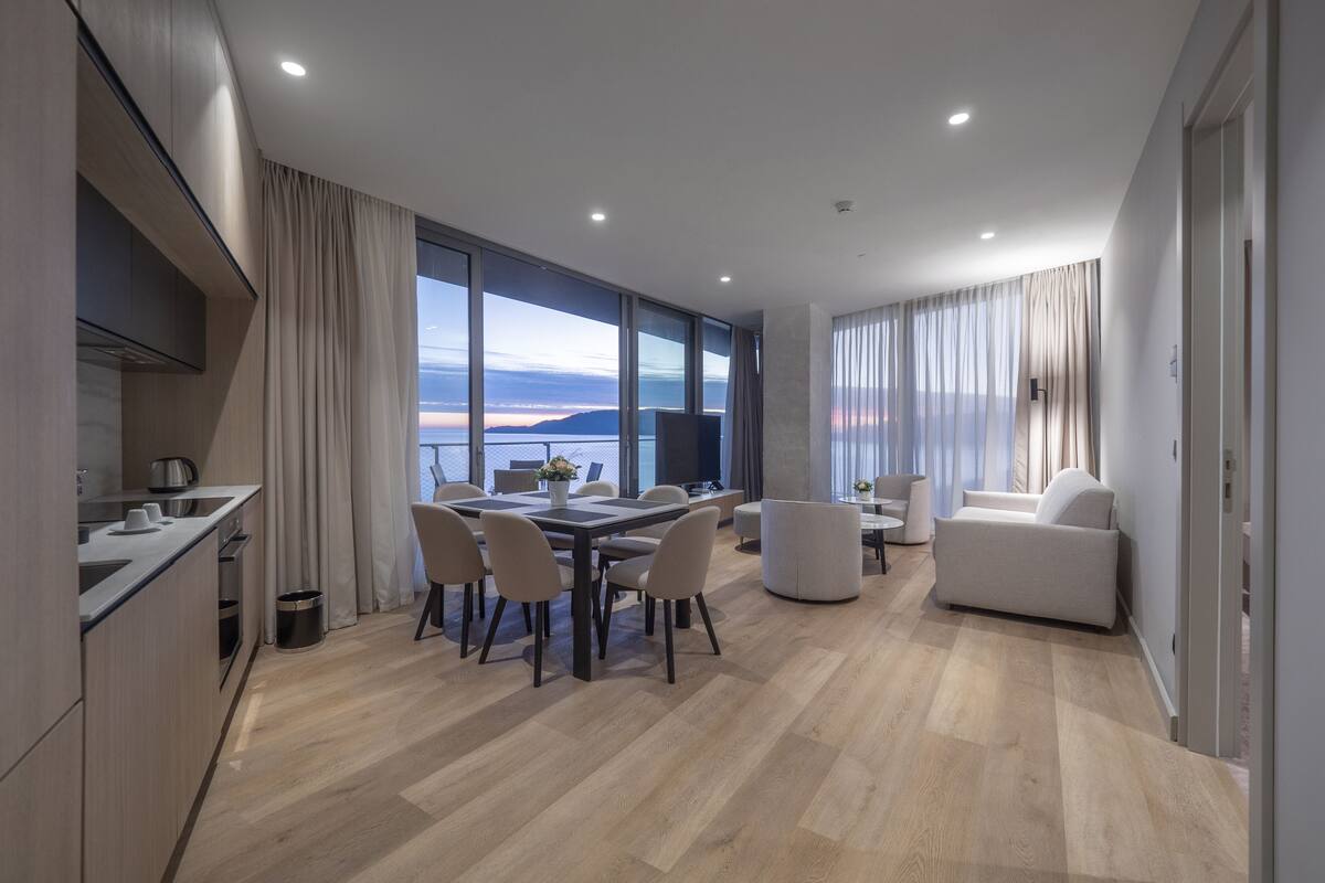 A modern living room with a dining area, large windows, and a view of the sea.