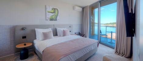 Une chambre d’hôtel moderne avec un grand lit, une petite table ronde, une chaise, une télévision et un balcon avec vue sur la mer.
