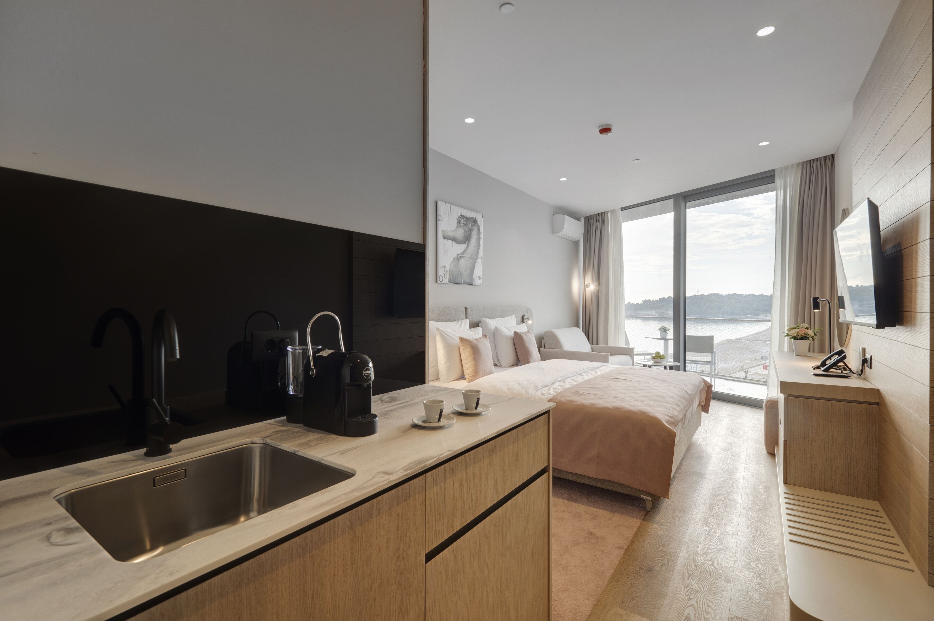 Ein modernes Hotelzimmer mit einem großen Bett, einer Küchenzeile mit Spüle und Kaffeemaschine, einem Flachbildfernseher und Blick auf den Strand.