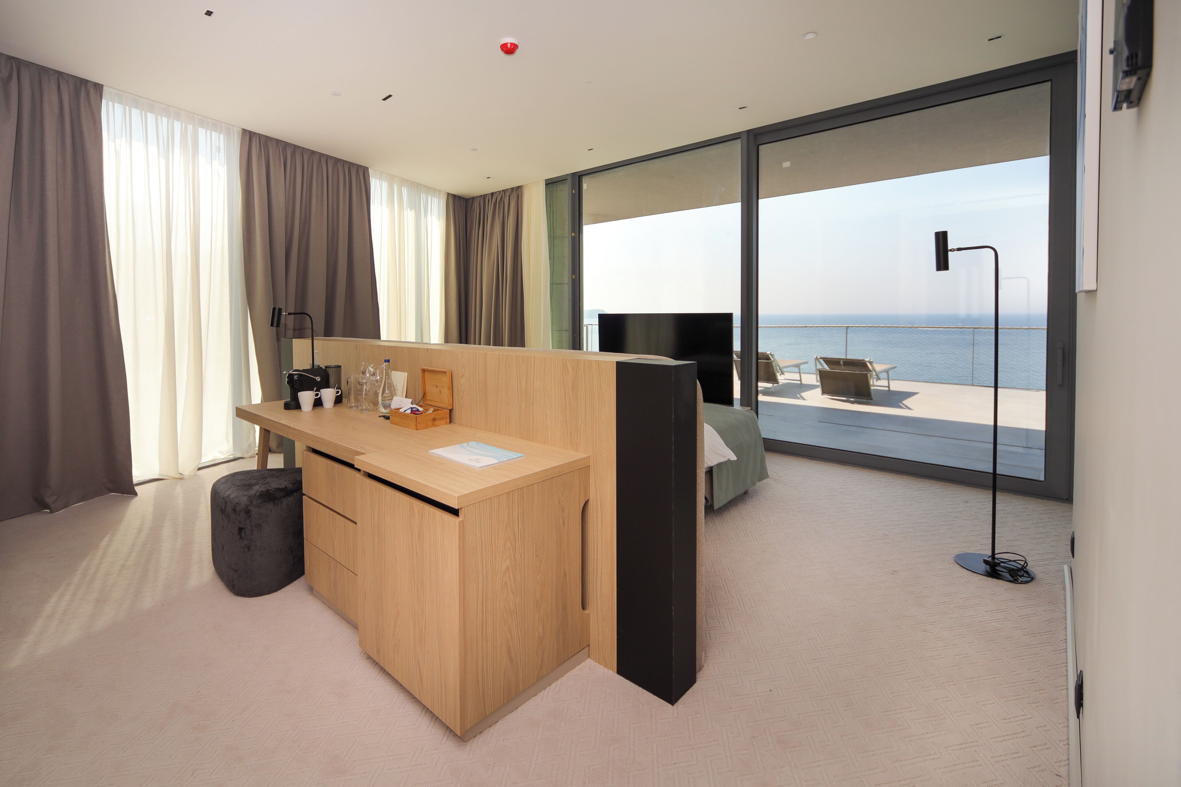 A modern hotel room with a wooden desk, a flat-screen TV, a large window with a view of the sea, and a balcony with outdoor seating.