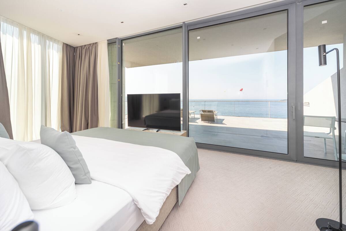 A modern hotel room with a large bed, a TV, and a sliding glass door leading to a balcony with a view of the sea.