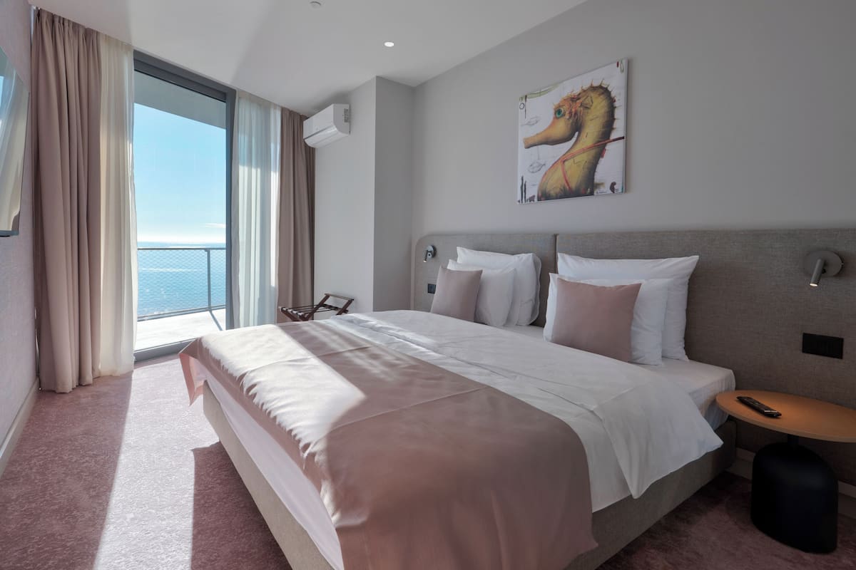 A hotel room with a large bed, a view of the sea, a painting on the wall, and a small table.