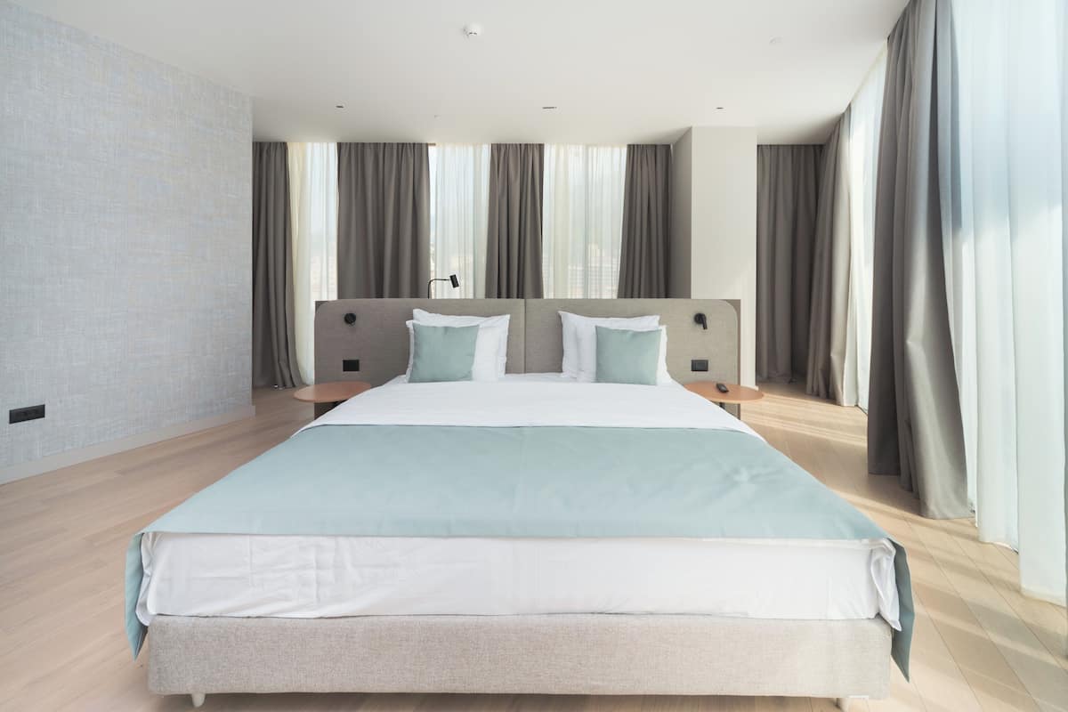 A modern hotel room with a large bed, neutral color scheme, and large windows with curtains.