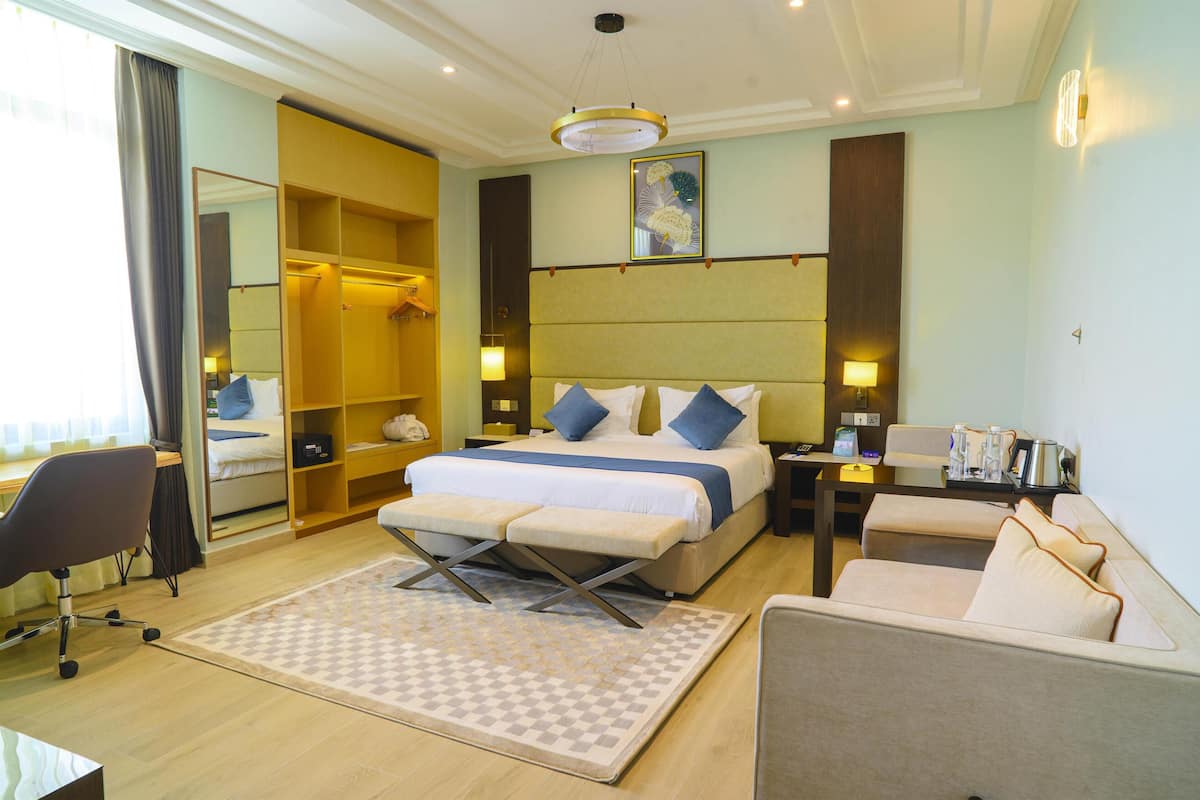 A hotel room with a large bed, bedside tables, a desk, a chair, a sofa, a mirror, and a wardrobe.