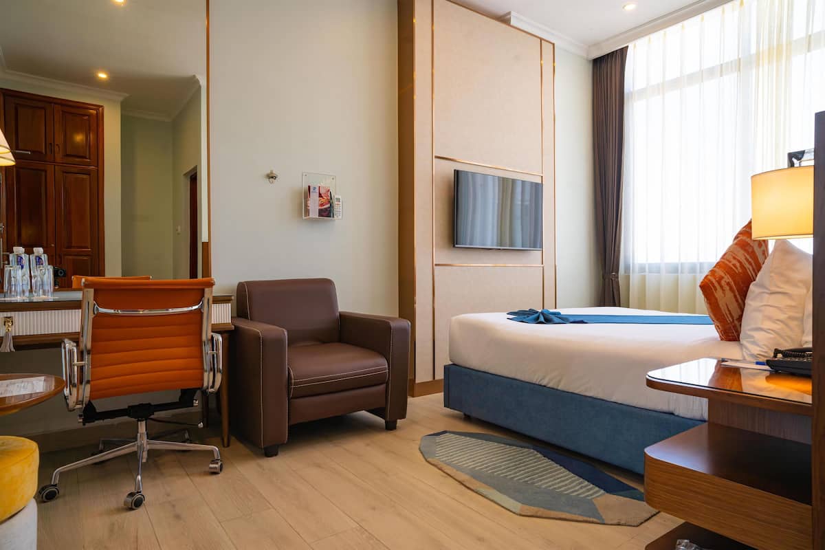 A hotel room with a bed, a desk with a lamp, a chair, a TV, and a bathroom area with cabinets and a mirror.