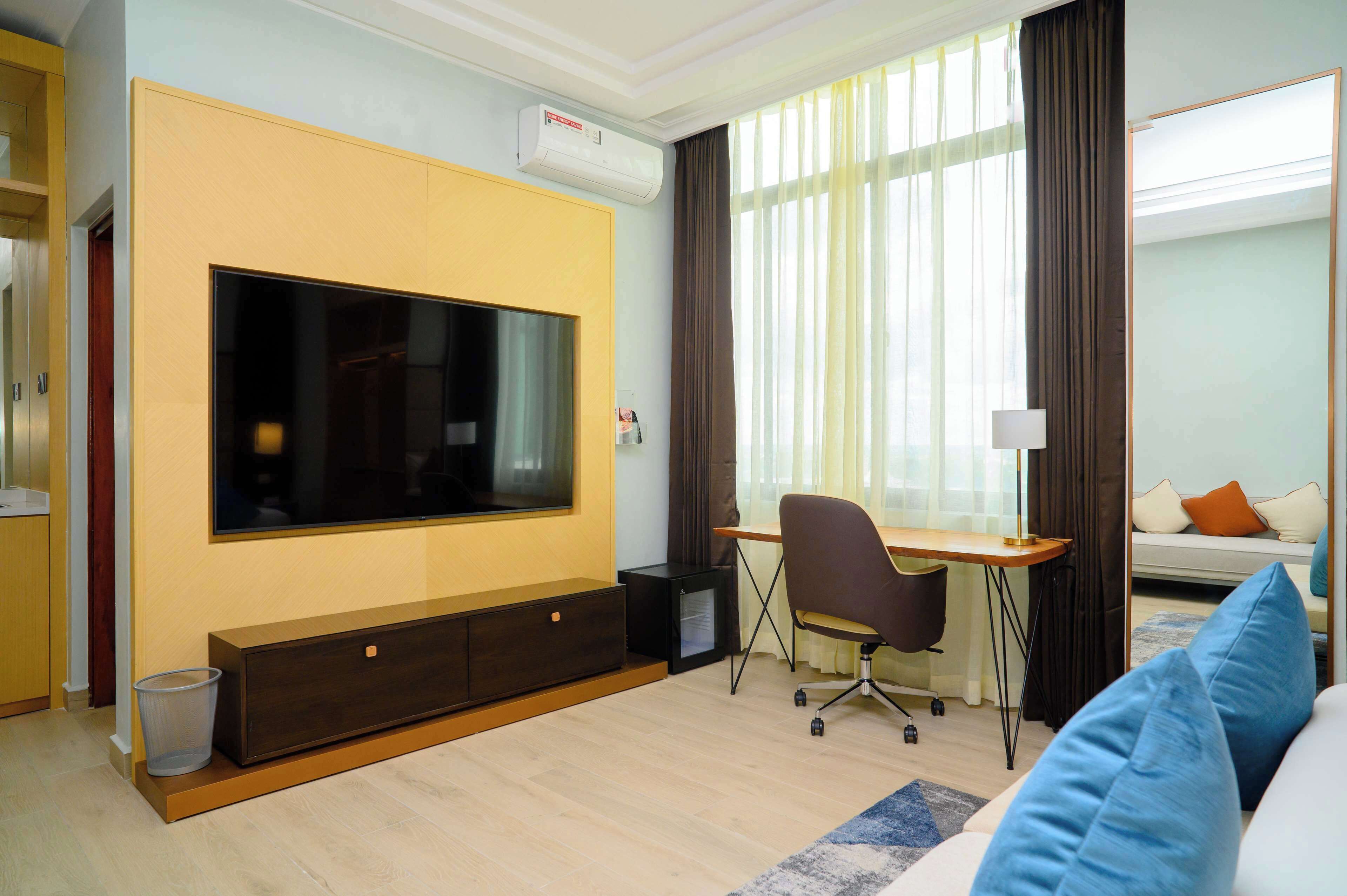 A modern hotel room with a large flat-screen TV, a desk with a chair, a sofa, and a bed with pillows.