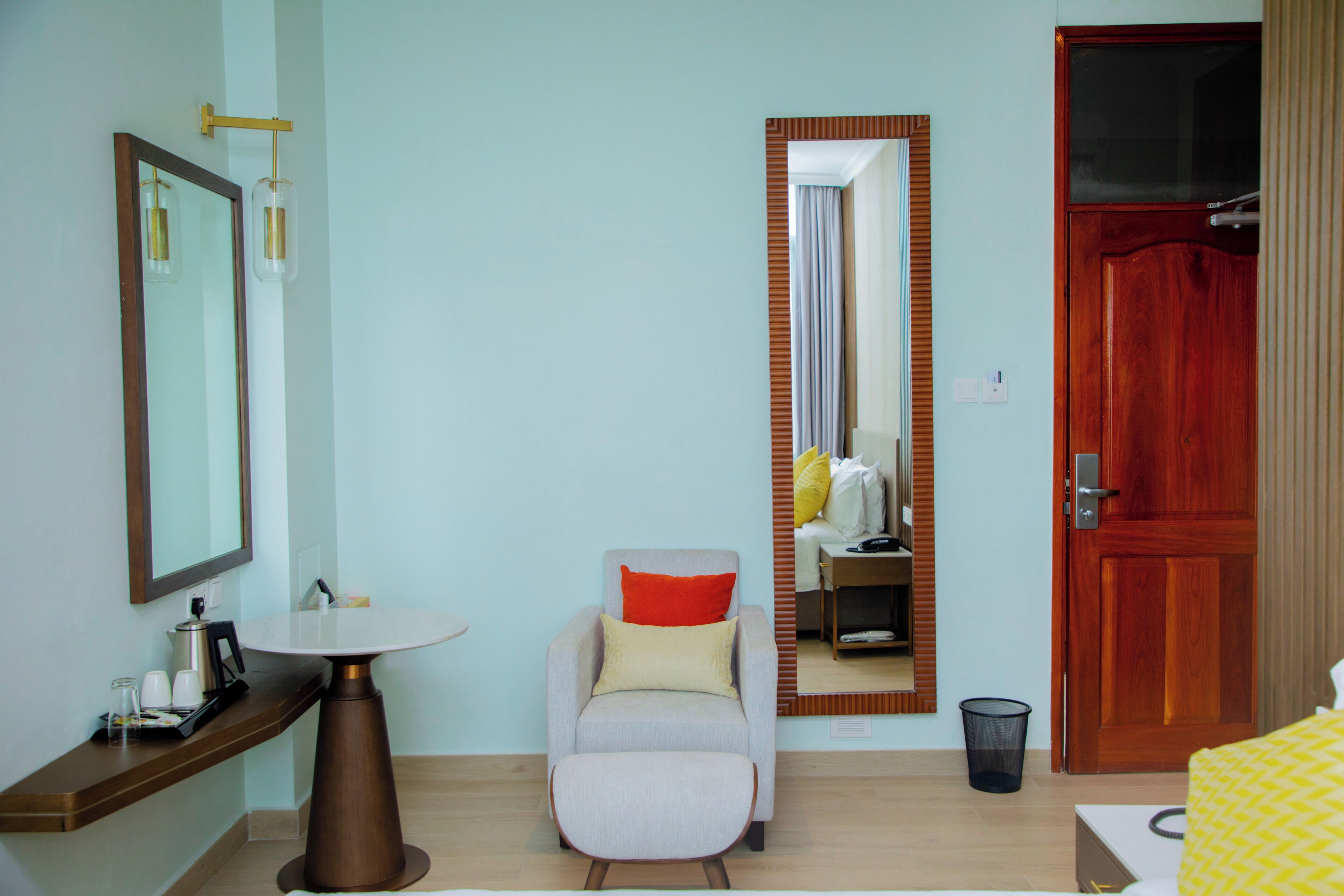 A hotel room with a round table, a chair with a red cushion, a large mirror, a wooden door, and a small trash bin.
