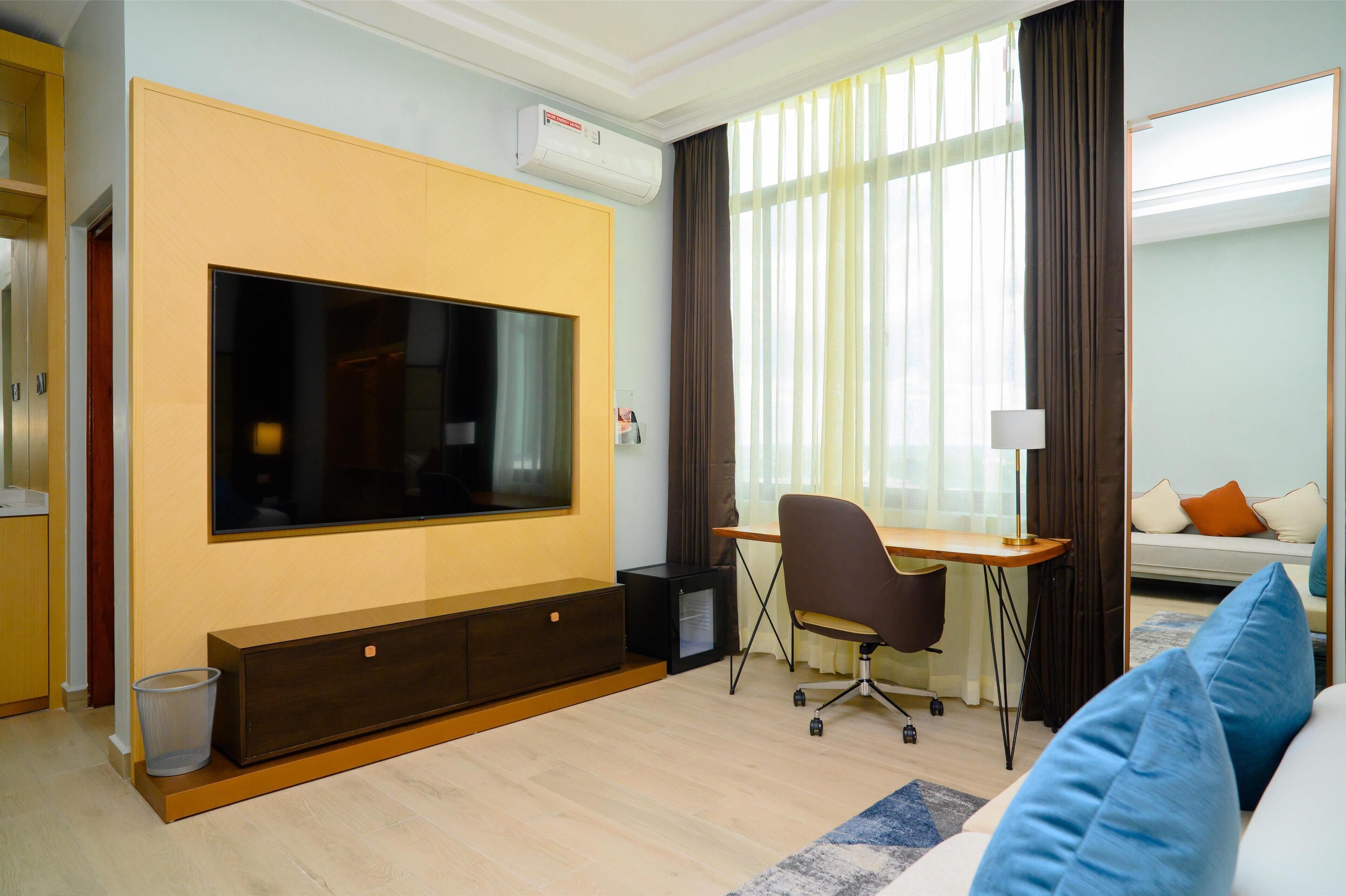 A modern hotel room with a large flat-screen TV, a desk with a chair, a sofa, and a window with curtains.