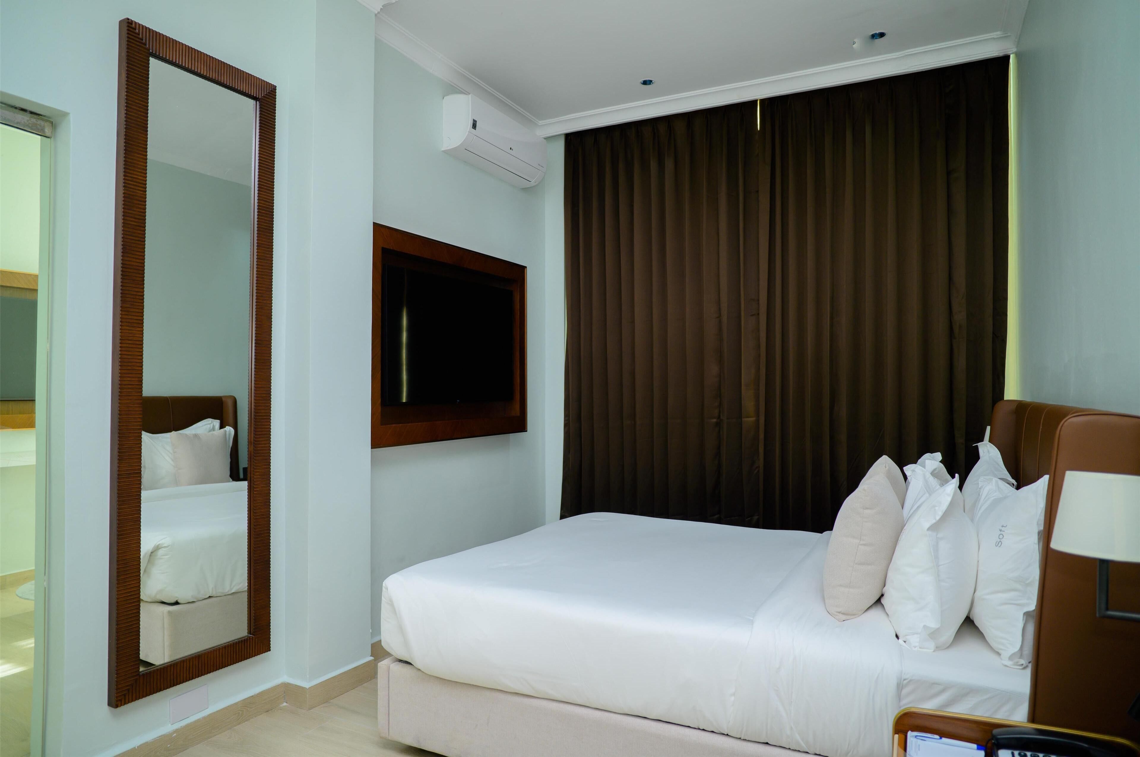Una habitación de hotel con cama, un espejo grande, un televisor de pantalla plana y una ventana con cortinas marrones.