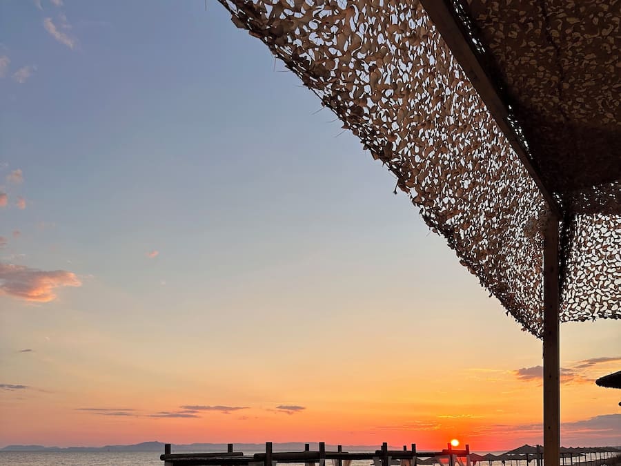 A beachside seating area with wicker chairs and cushions, a wooden pergola, and a view of the sunset over the ocean.
