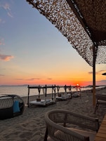 Un área de estar frente a la playa con sillas de mimbre y cojines, una pérgola de madera y vistas al atardecer sobre el océano.