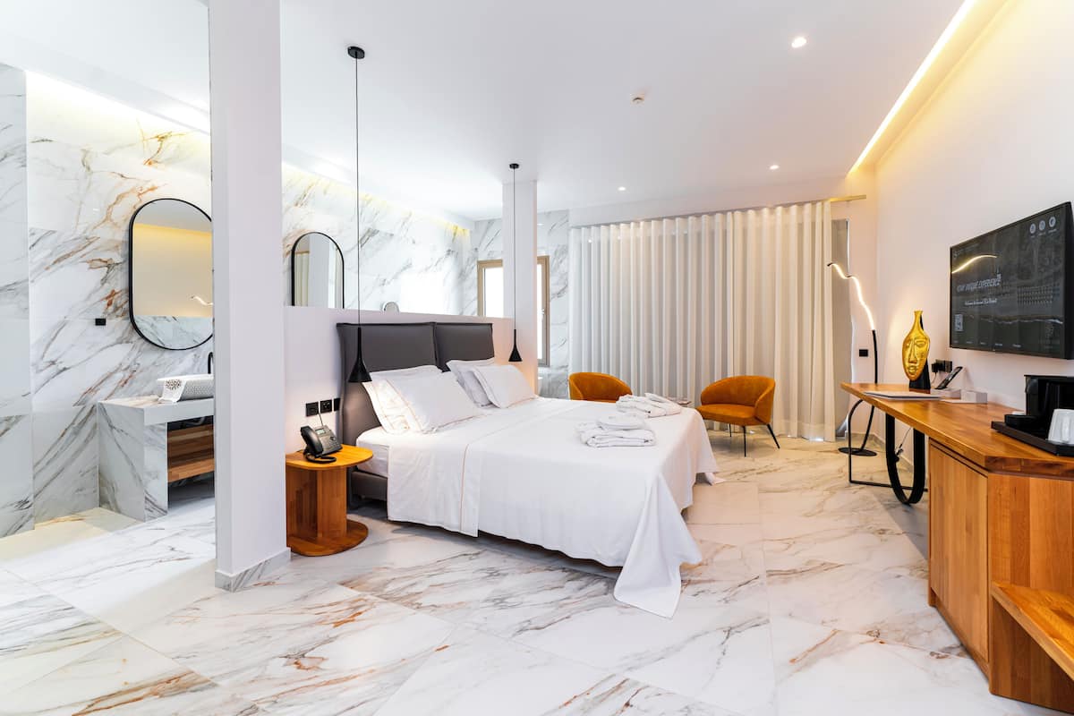 Moderni hotellihuone, jossa on marmorilattia, valkoisilla lakanoilla varustettu sänky, puinen työpöytä, tuoli, televisio ja suuri ikkuna, jossa on verhot.