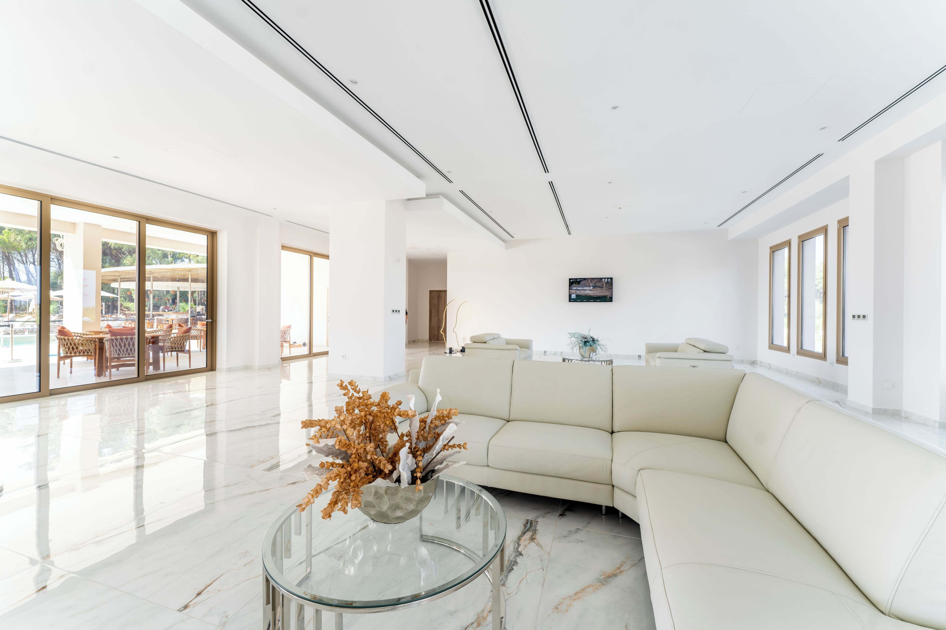 A spacious living area with a white sofa, a glass coffee table with a decorative plant, and a flat-screen TV mounted on the wall.