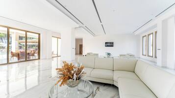 A spacious living area with a white sofa, a glass coffee table with a decorative plant, and a flat-screen TV mounted on the wall.