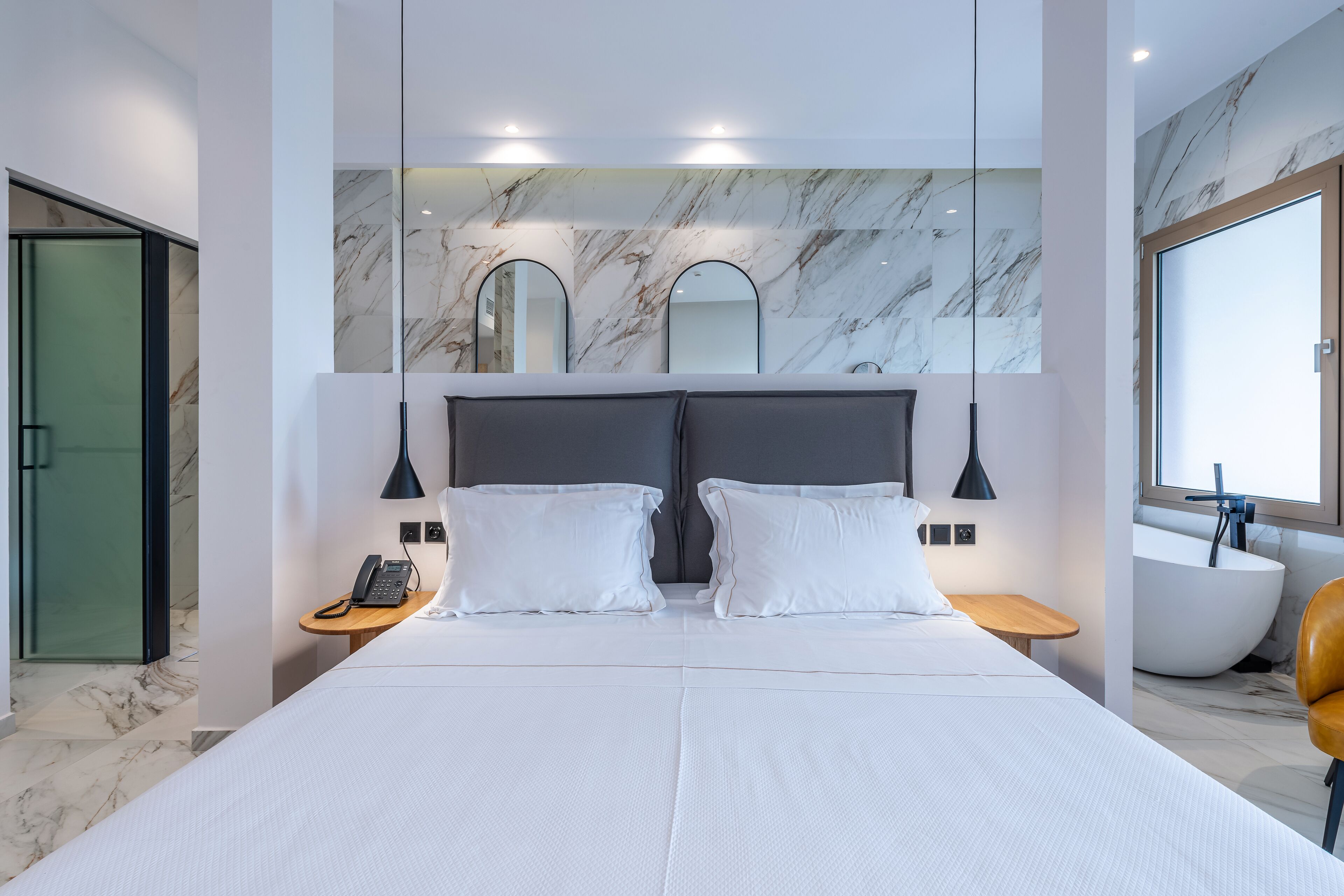 Une chambre d’hôtel moderne dotée d’un grand lit, de deux tables de chevet avec lampes, d’une baignoire intégrée et d’un mur en marbre.