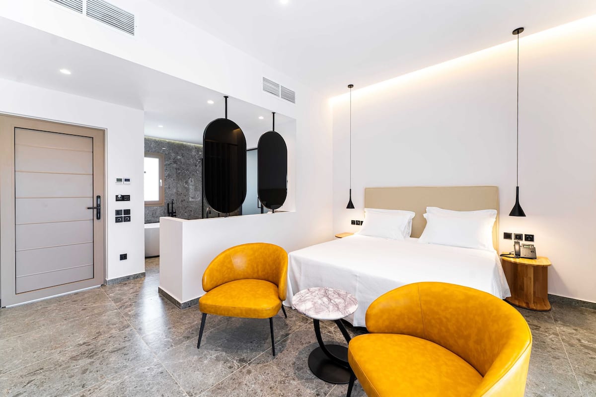 Moderni hotellihuone, jossa on suuri sänky, kaksi keltaista nojatuolia, marmoripintainen pöytä, puinen sivupöytä ja suuri peili.