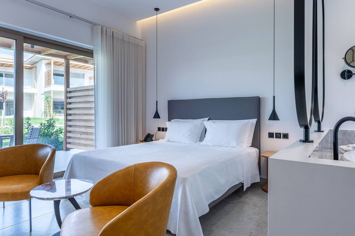 Moderni hotellihuone, jossa on suuri sänky, kaksi tuolia, pieni pöytä, kylpyhuone, jossa on pesuallas, ja näkymä ulos.