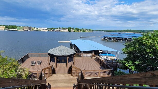 Eine Holzterrasse führt zu einem Pavillon mit Blick auf einen See, Boote und Gebäude in der Ferne.