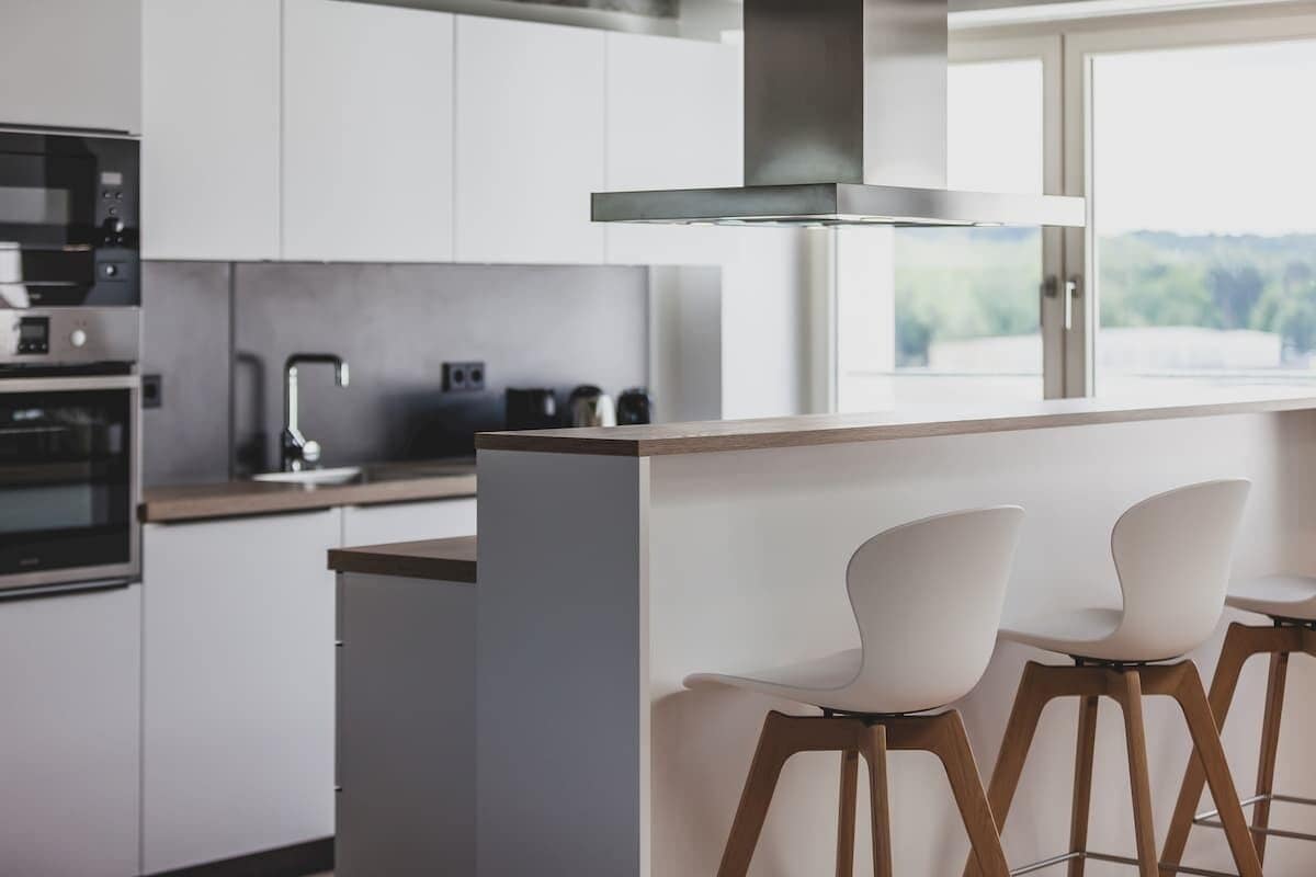 A modern kitchen with white cabinets, a stainless steel range hood, and a bar area with white bar stools.