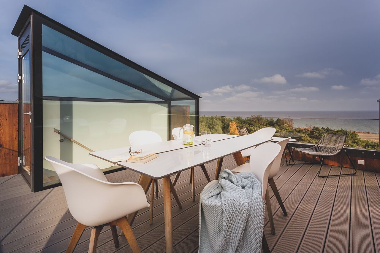 A modern outdoor dining area with a glass enclosure, wooden deck, and a view of the sea.