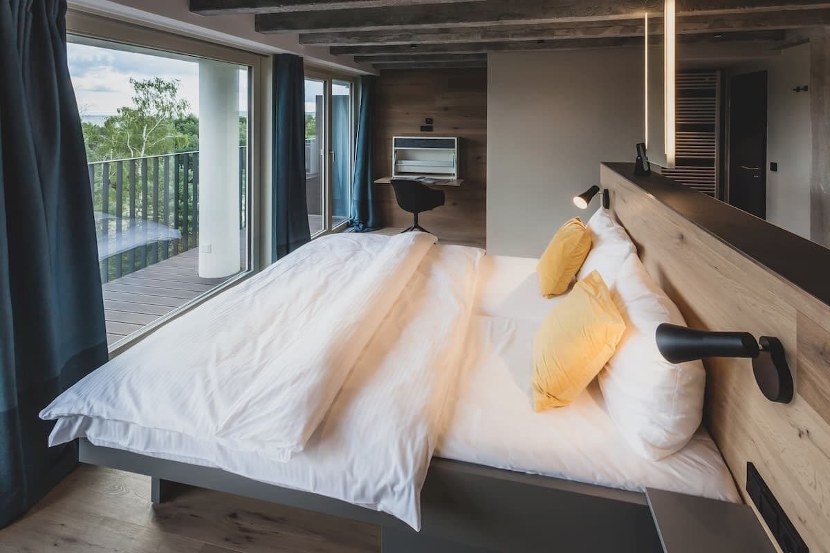 A modern bedroom with a large bed, a desk, and a balcony with a view.