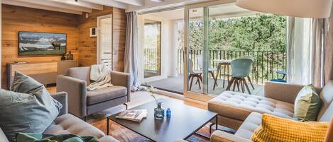 A modern living room with a sofa, armchairs, and a coffee table. There is a television mounted on the wall and a sliding glass door leading to an outdoor area with a table and chairs.