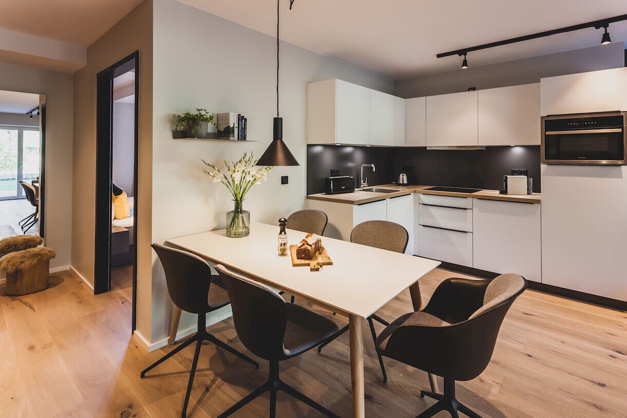 Une cuisine moderne avec des meubles blancs, un plan de travail noir et un four encastré. On y trouve une table à manger avec quatre chaises et un vase de fleurs.
