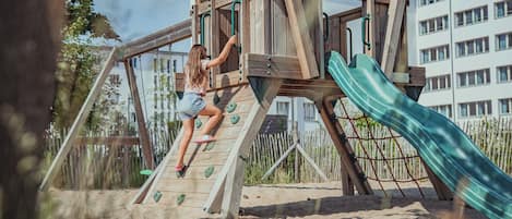 A wooden play structure with a slide and climbing wall, surrounded by sand and trees.
