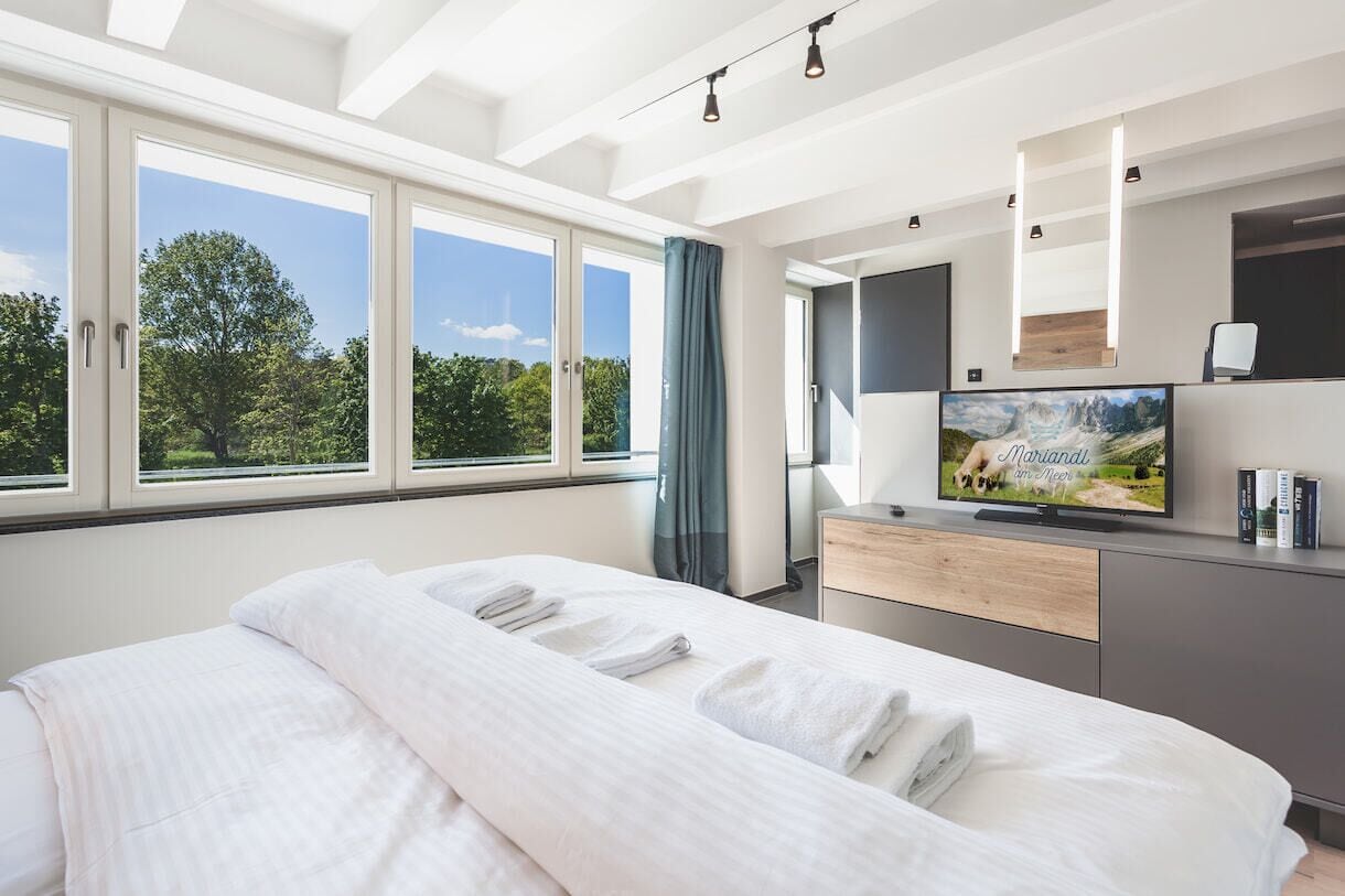 Ein modernes Hotelzimmer mit einem großen Bett, einem Fernseher und Blick auf Bäume durch die Fenster.