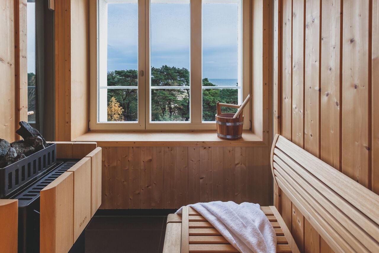 A wooden sauna room with a bench, a bucket, and a window overlooking a natural landscape.