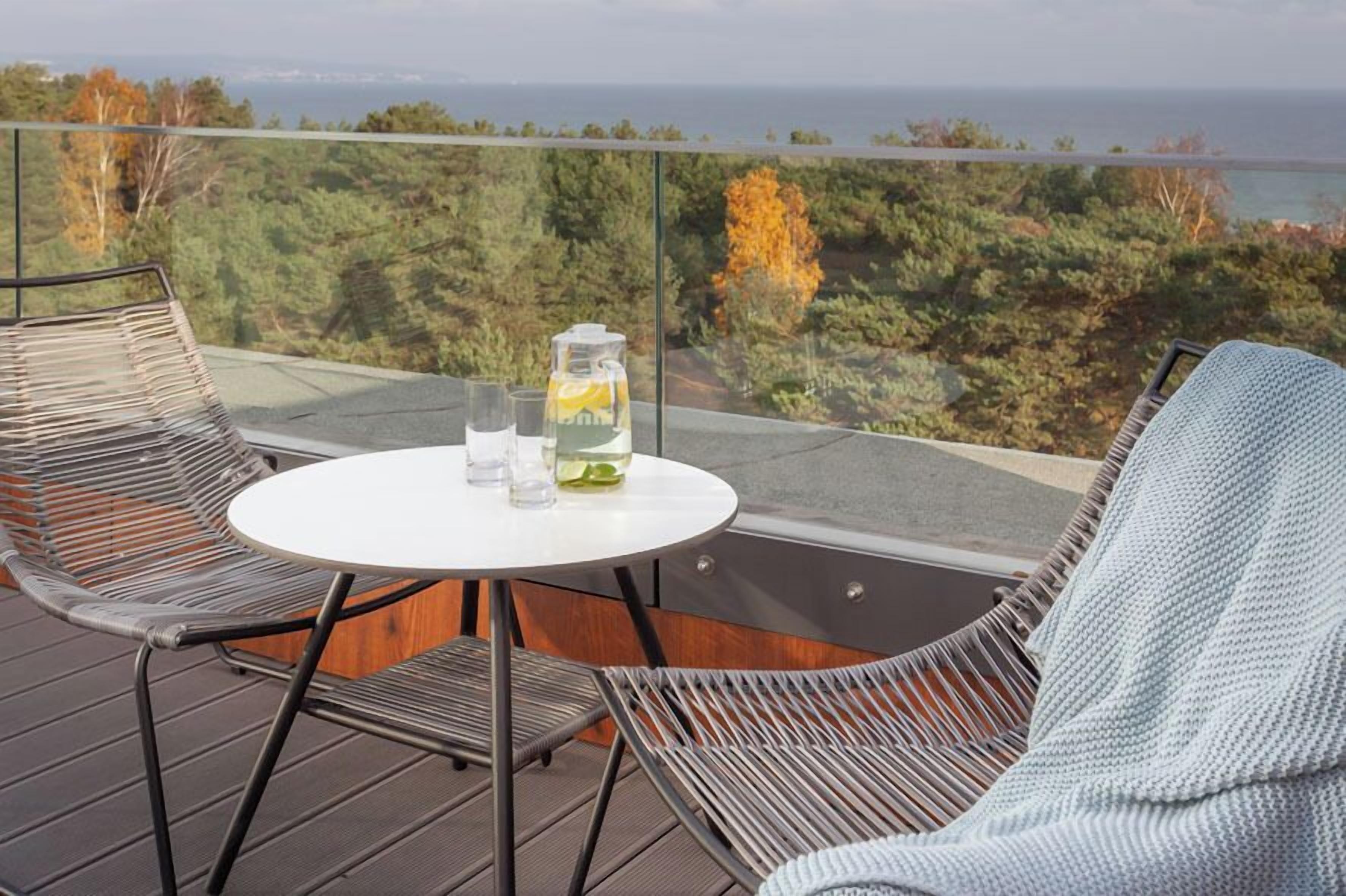 Ein Balkon mit einem runden Tisch und zwei Korbstühlen, der einen Blick auf bewaldetes Gelände und das Meer bietet.