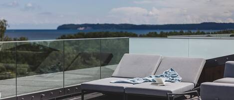 Et moderne udendørs loungeområde med gelænder i glas, grå loungesæder og udsigt til en kystlinje.