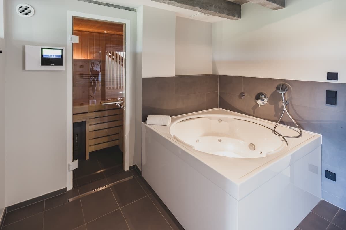 A modern bathroom with a large bathtub, a shower, and a toilet. There is a control panel on the wall and a wooden door leading to another room.