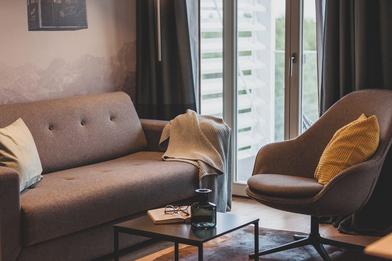 A modern living room with a sofa and armchair, a coffee table with a book and glasses, and a view through large windows.