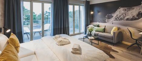 A modern hotel room with a large bed, a sofa, a small table, and a view of the outdoors.