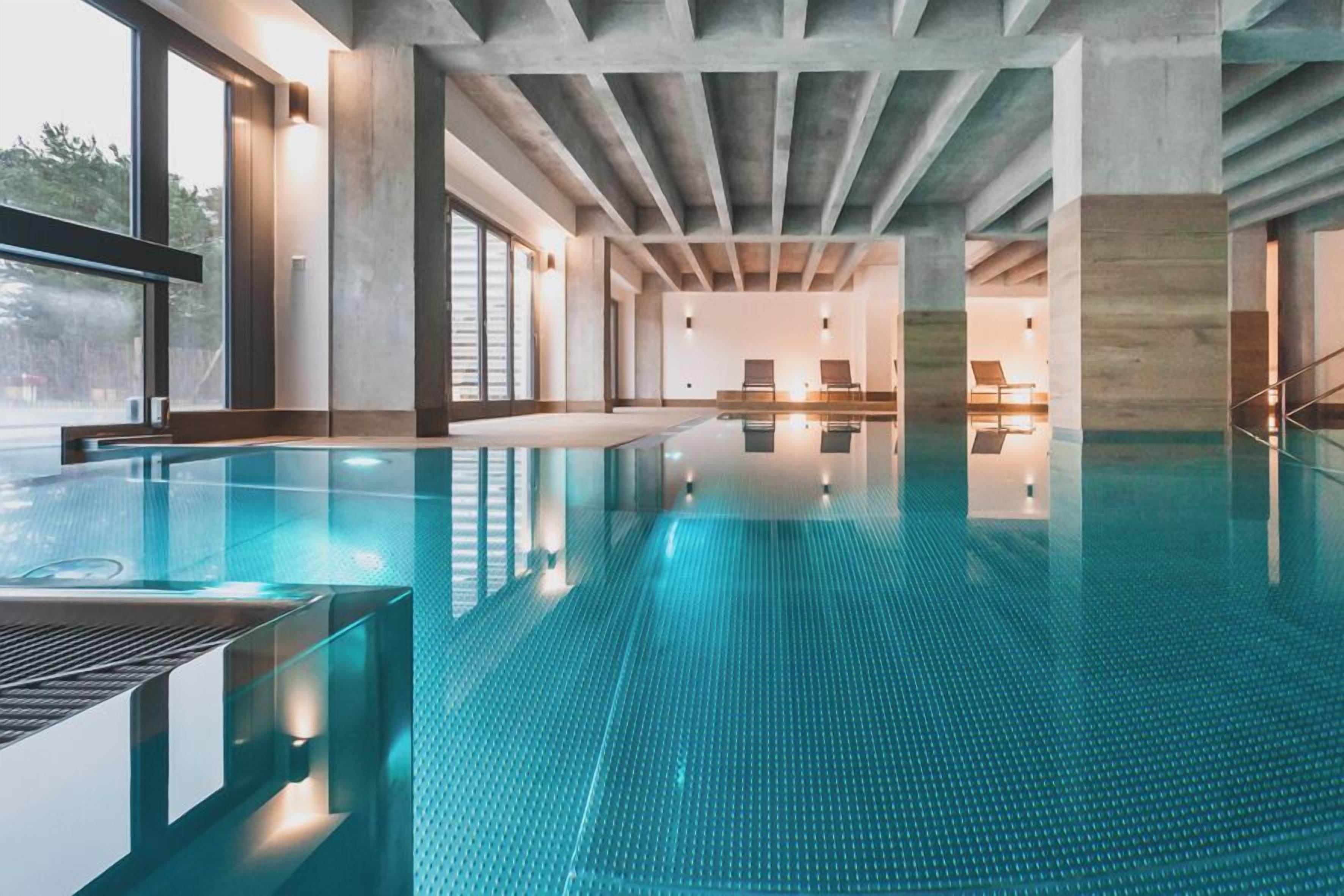An indoor swimming pool with a clear blue tiled floor, large windows, and a modern architectural design with exposed concrete beams.