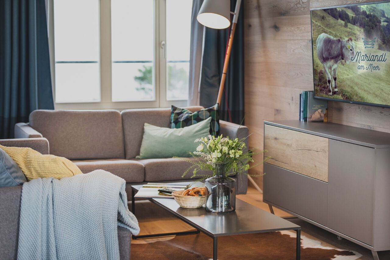 A modern living room with a grey sofa, a coffee table with a vase of flowers, a television on the wall, and a bookshelf.