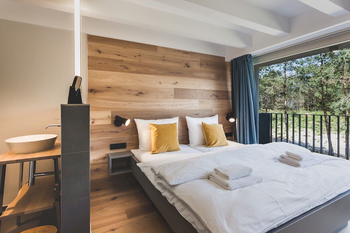 A modern bedroom with a wooden headboard, a large bed with white linens, a bedside table with a white bowl, and a view of the outdoors through a balcony.