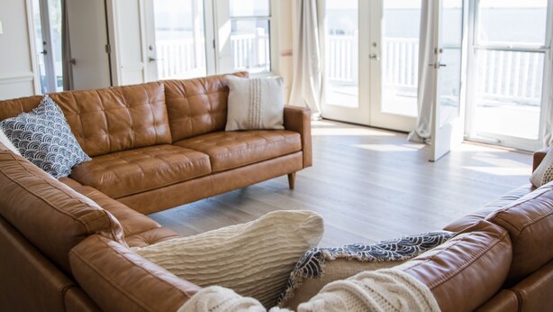A spacious living room with a brown leather sofa, a white knitted throw, and a ceiling fan.