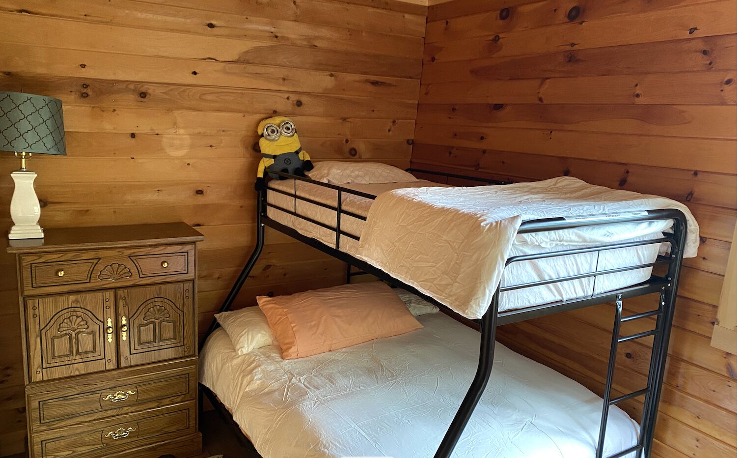 A bunk bed with a Minion toy on the top bunk, a wooden dresser with drawers, and a lamp with a patterned lampshade.
