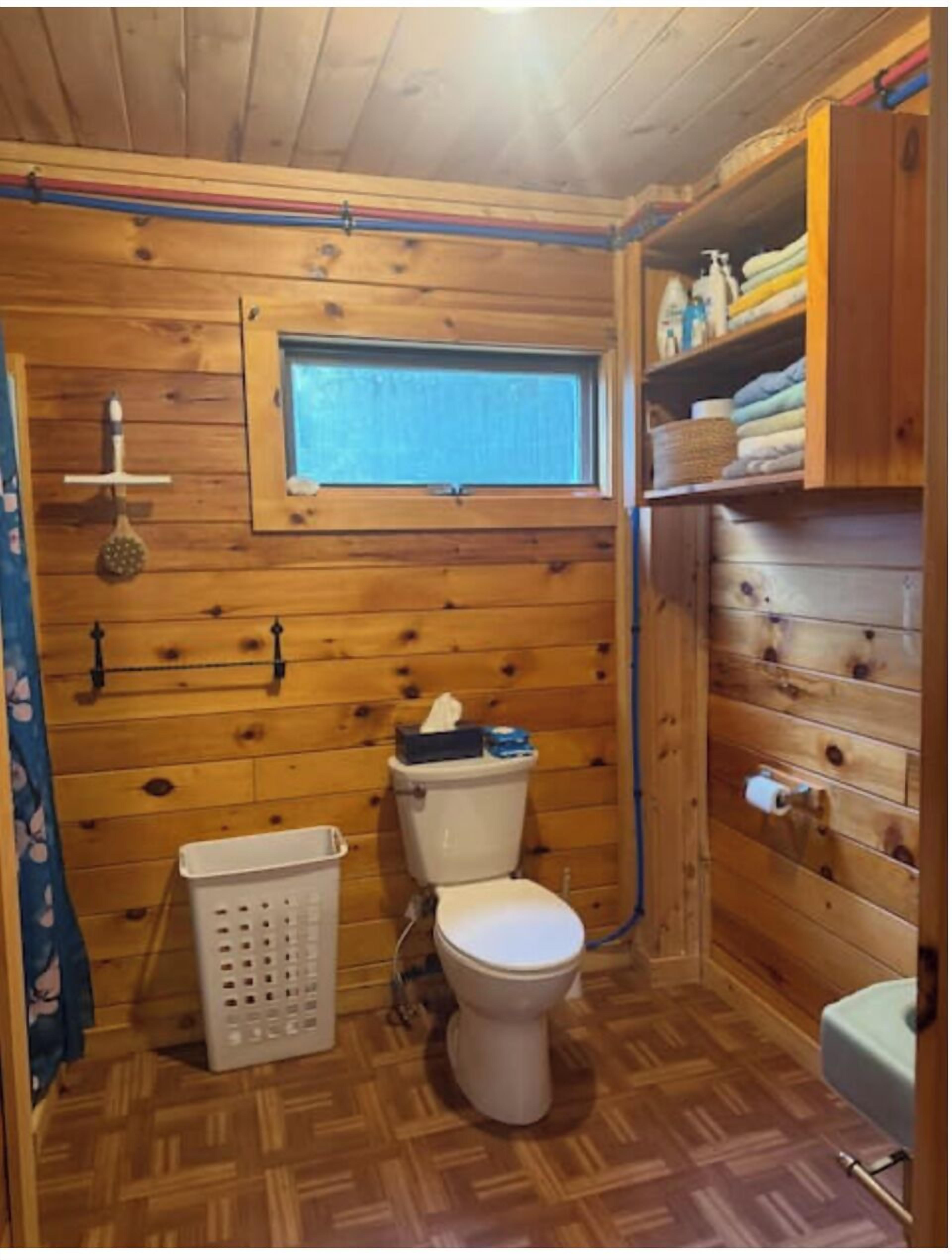 A wooden-paneled bathroom with a toilet, a window, and a shelf with folded towels.