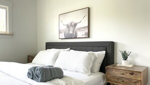 Un dormitorio con cama, almohadas, mesita de noche con una planta y un cuadro en la pared.