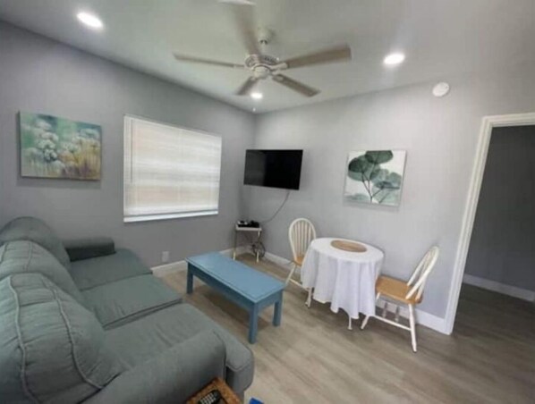 A living room with a grey sofa, a blue coffee table, a white dining table with chairs, a ceiling fan, and artwork on the walls.