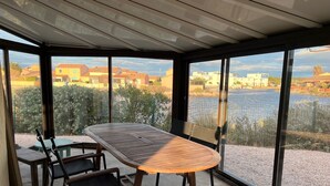A sunlit patio with a wooden table and chairs, large windows offering a view of a body of water and surrounding buildings. - Barcarès, ile des Pêcheurs, Mas Bord de Marina (Le Barcarès)